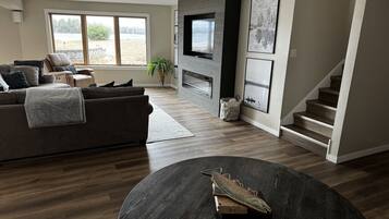 A modern living room with a dark wood floor, a brown leather sofa, a flat-screen TV mounted on a wooden wall, and a staircase leading to another level.