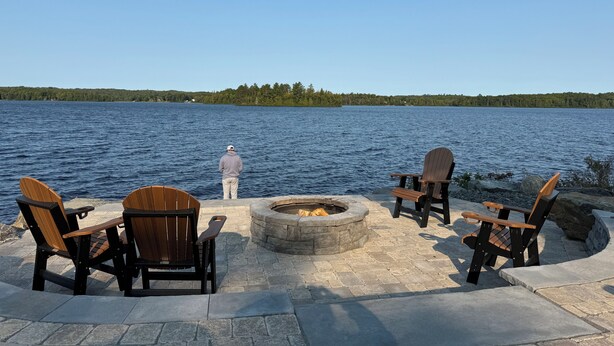 Un espacio de descanso junto al lago con sillas Adirondack y un hogar para fuego, con vista a un lago tranquilo bordeado por un bosque.