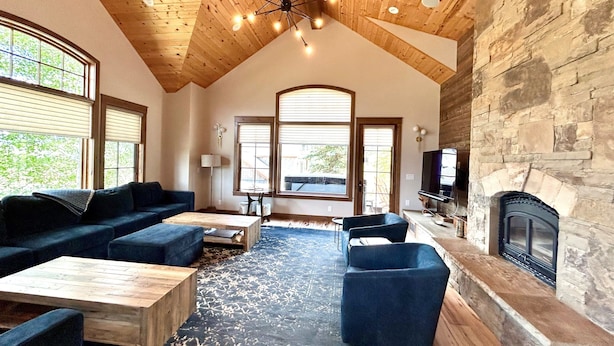 A spacious living room with a high ceiling, wooden beams, a stone fireplace, and large windows.