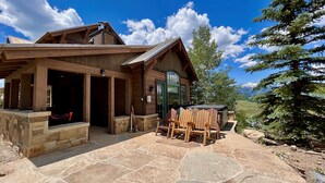 A wooden lodge with a stone patio, a hot tub, and a view of the mountains.
