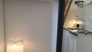 A room with a small table, a lamp, and a large mirror reflecting a kitchen area. - Charming studio in the Marais (Paris)
