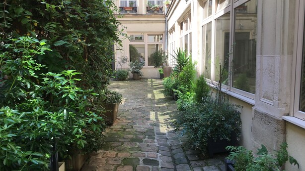 A narrow stone pathway flanked by lush greenery and potted plants, leading to a building with large windows and a balcony. - Charming studio in the Marais (Paris)