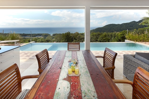 Un espace repas offrant une vue sur l’océan et les collines, une piscine et du mobilier d’extérieur.