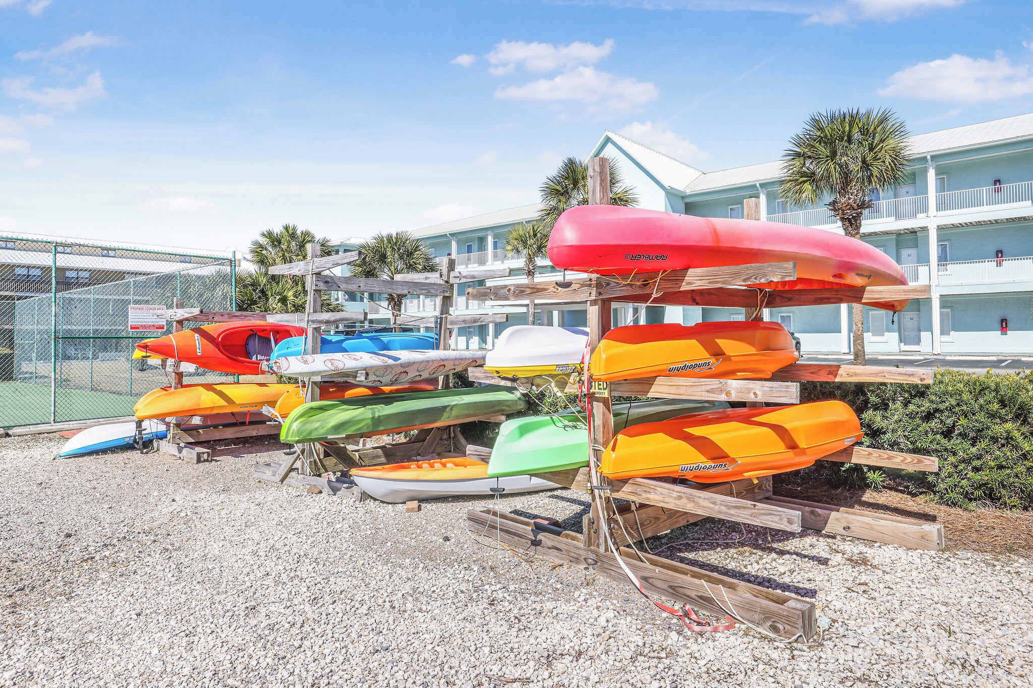 Varias kayaks y tablas de remo sobre estantes de madera cerca de un edificio con piscina y palmeras.