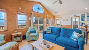 A cozy living room with a blue sofa, a matching armchair, and a wooden coffee table. There is a large window offering a view of the ocean.