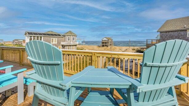 A deck with two light blue Adirondack chairs and a small table, overlooking a beach with houses and the ocean.