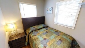 A bedroom with a bed, a nightstand, a lamp, and two windows with blinds.