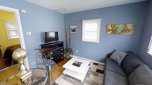 A living room with a sofa, a coffee table, a TV, a dining table with chairs, and artwork on the walls.