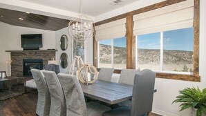 A dining area with a wooden table and grey upholstered chairs, a stone fireplace, and a large window with a view of a desert landscape.