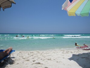 Une plage à l’eau d’un bleu cristallin, où l’on peut se baigner et faire de la planche à voile, et une rive sablonneuse parsemée de chaises longues et de parasols.