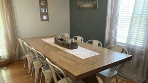 A dining area with a wooden table, white chairs, a hanging light fixture, and a window with curtains. - KY BOURBON TRAIL LAKEFRONT HOUSE WITH DOCK SLEEPS 8! Year round Hot tub! (Lawrenceburg)