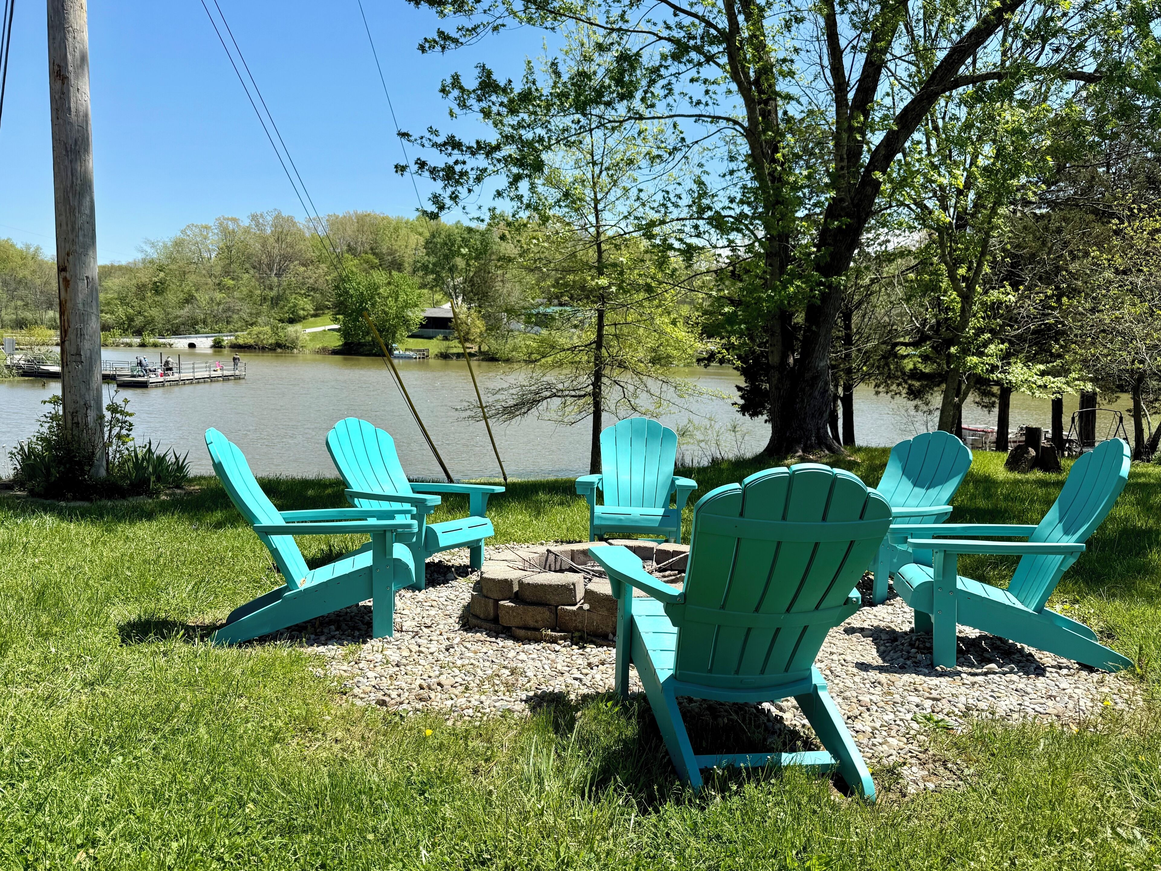 Un espacio de descanso junto al lago con sillas Adirondack turquesas, un brasero y vistas a un lago con barcos y un muelle al fondo.
