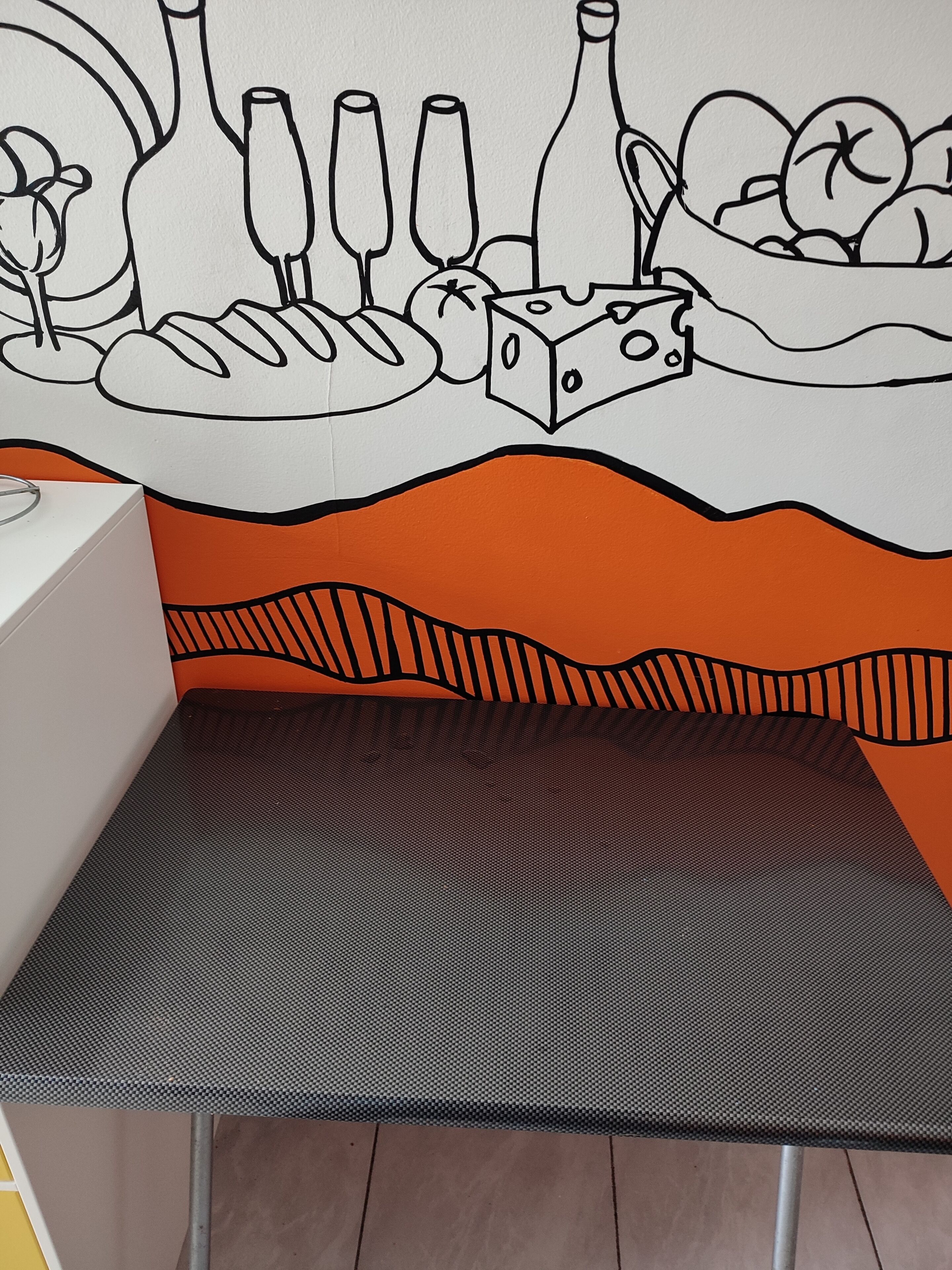 Un espace repas comprenant une table et des chaises, un mur orné d’une fresque représentant des aliments, et un sol à motifs.