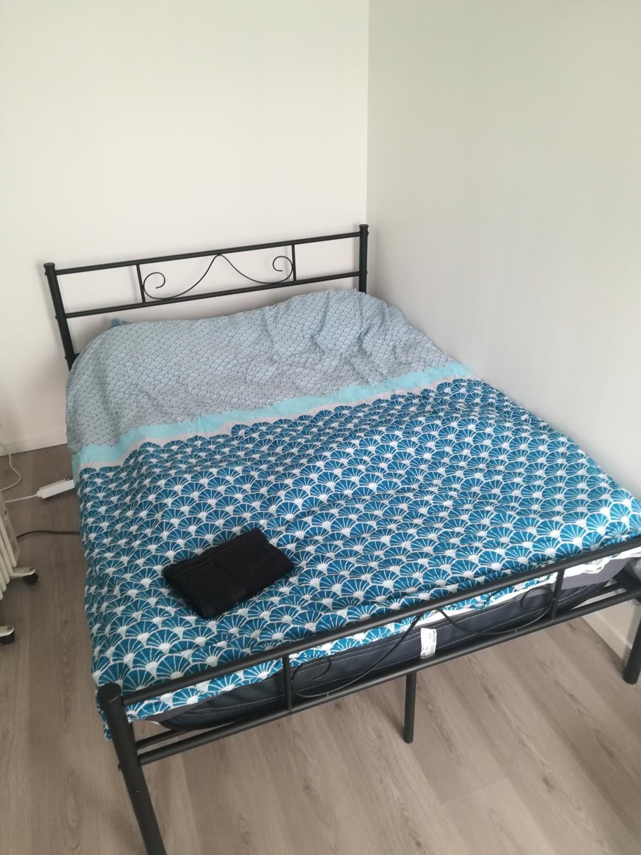 Una cama individual con estructura metálica, colcha azul y blanca estampada y una manta gris.