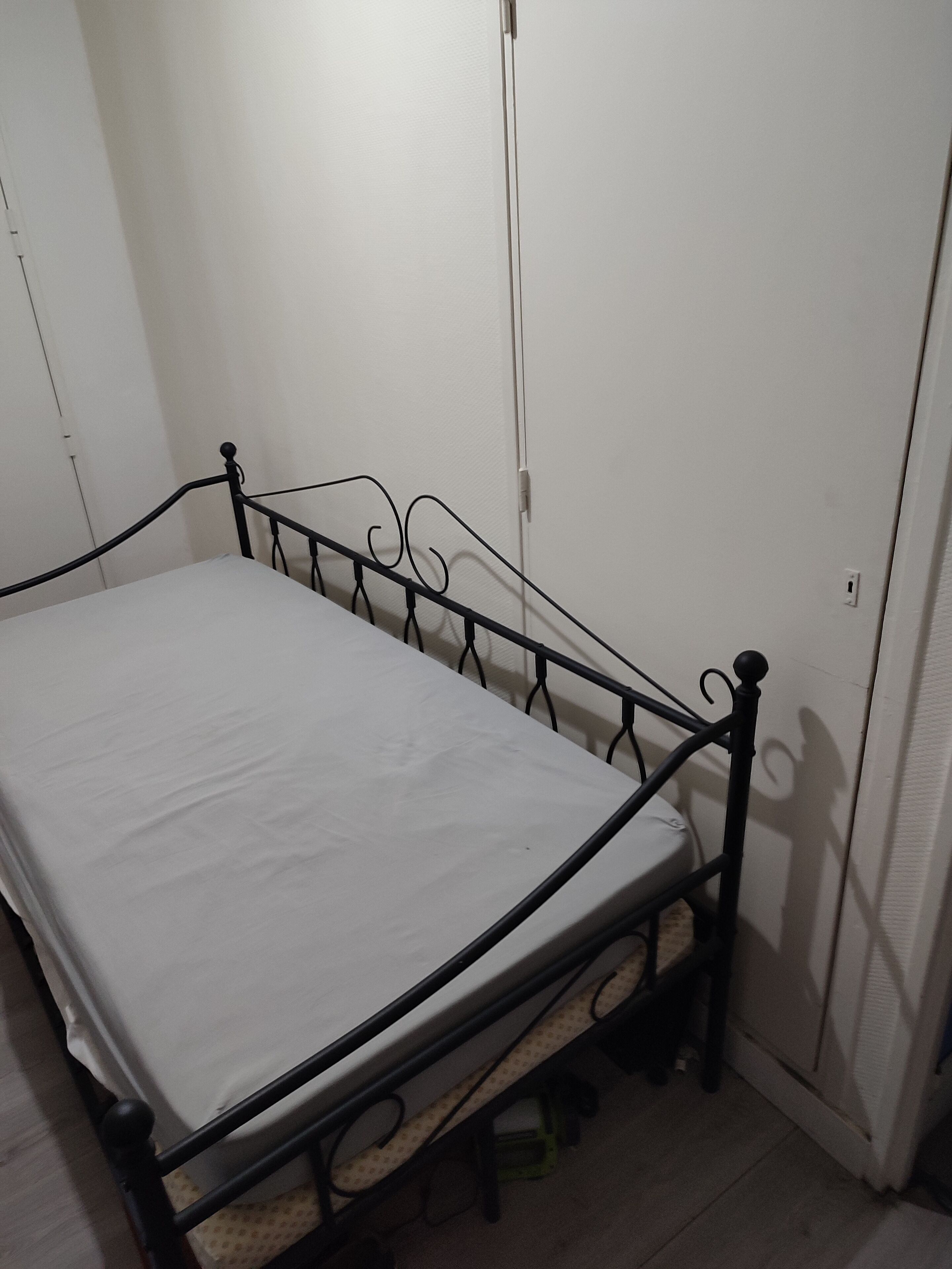 A single bed with a metal frame and a white mattress in a room with white walls and a wooden floor.