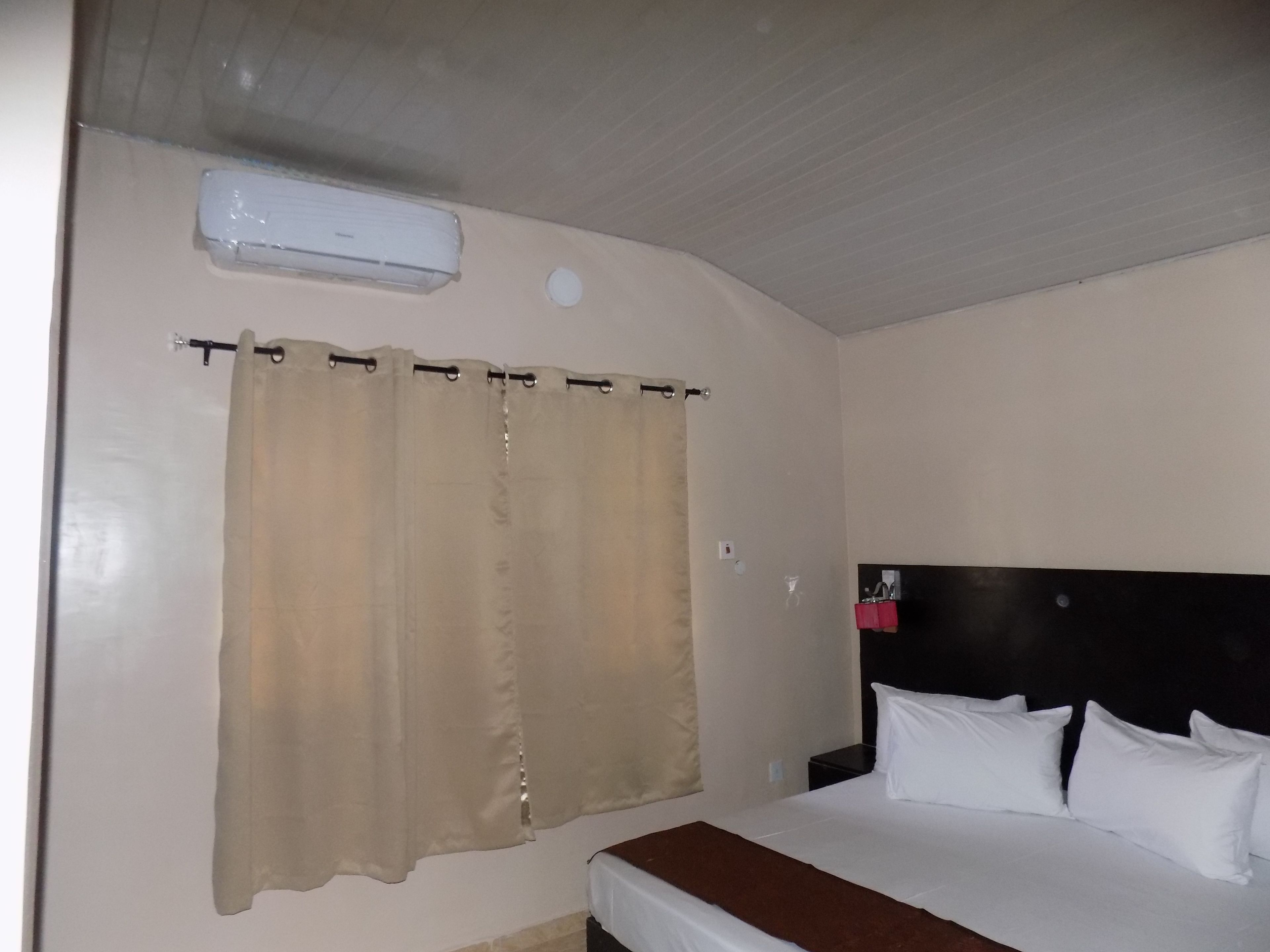 A hotel room with a bed, a headboard, a curtain, and an air conditioning unit.