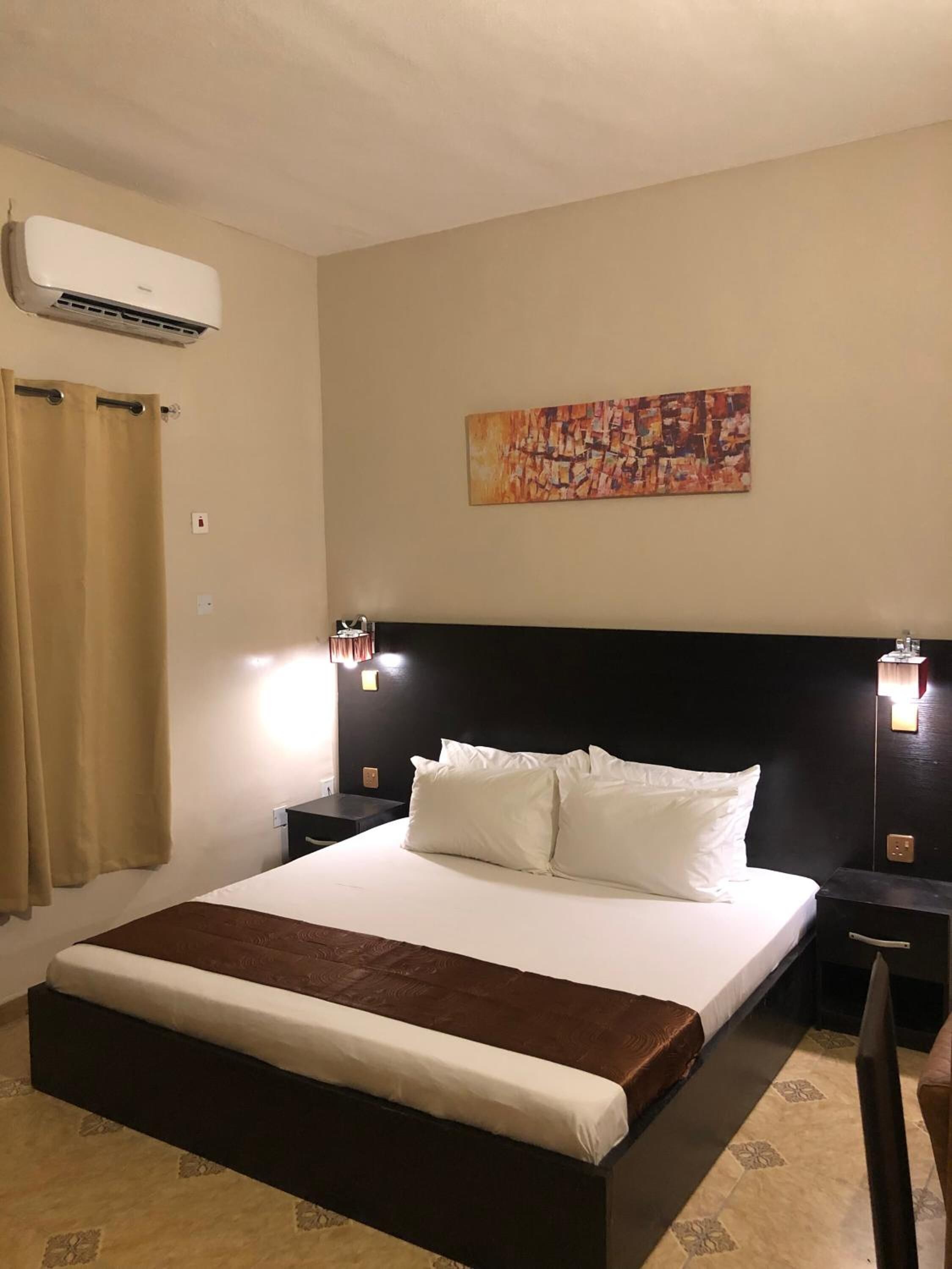 A hotel room with a large bed, a headboard, two bedside tables, an air conditioning unit, a painting on the wall, and a curtain.