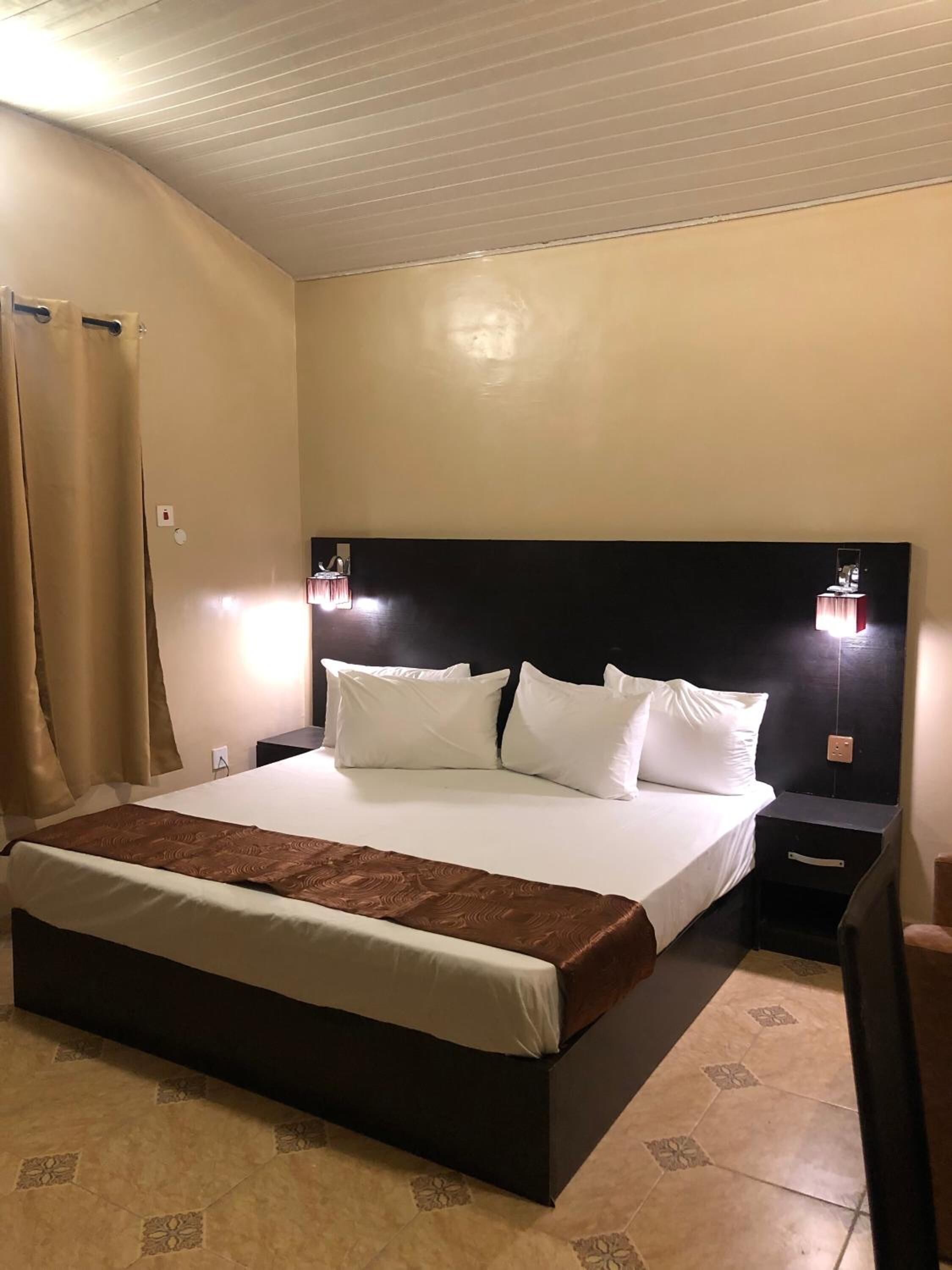 A hotel room with a large bed, two bedside tables, a headboard, and a curtain.