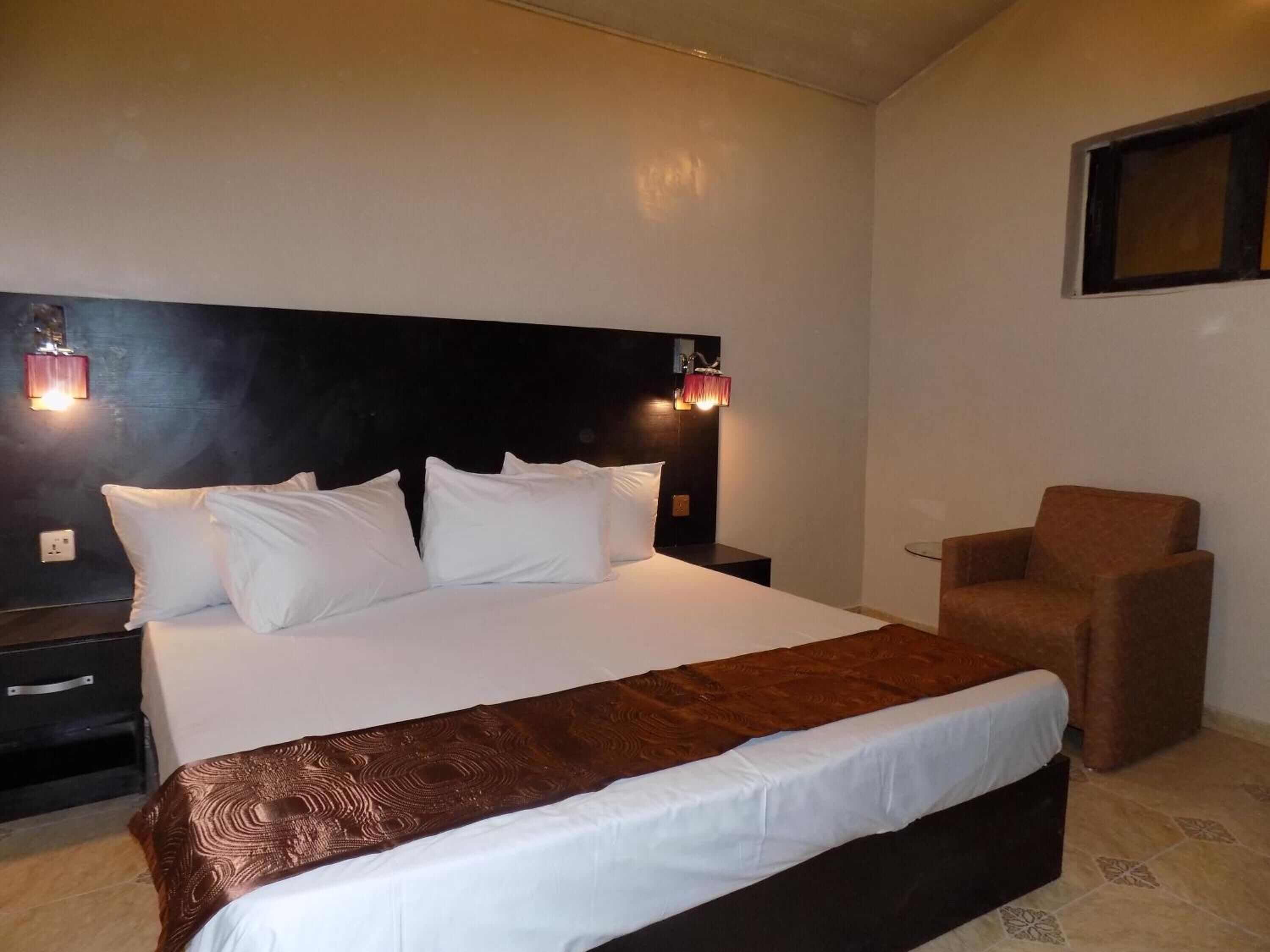 A hotel room with a large bed, a nightstand, a chair, and a wall-mounted light fixture.