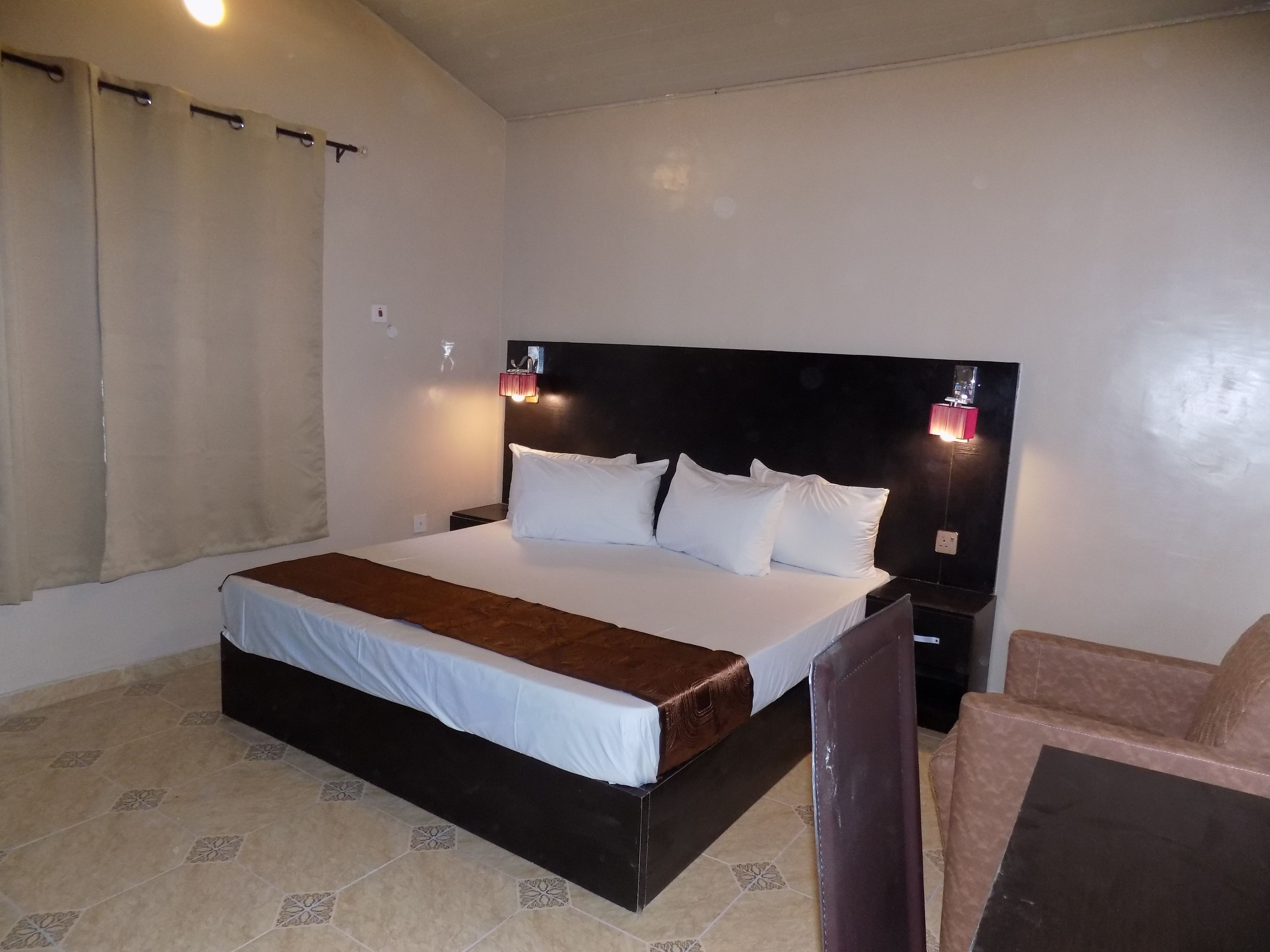 A hotel room with a large bed, a nightstand, a chair, and a wall-mounted lamp.