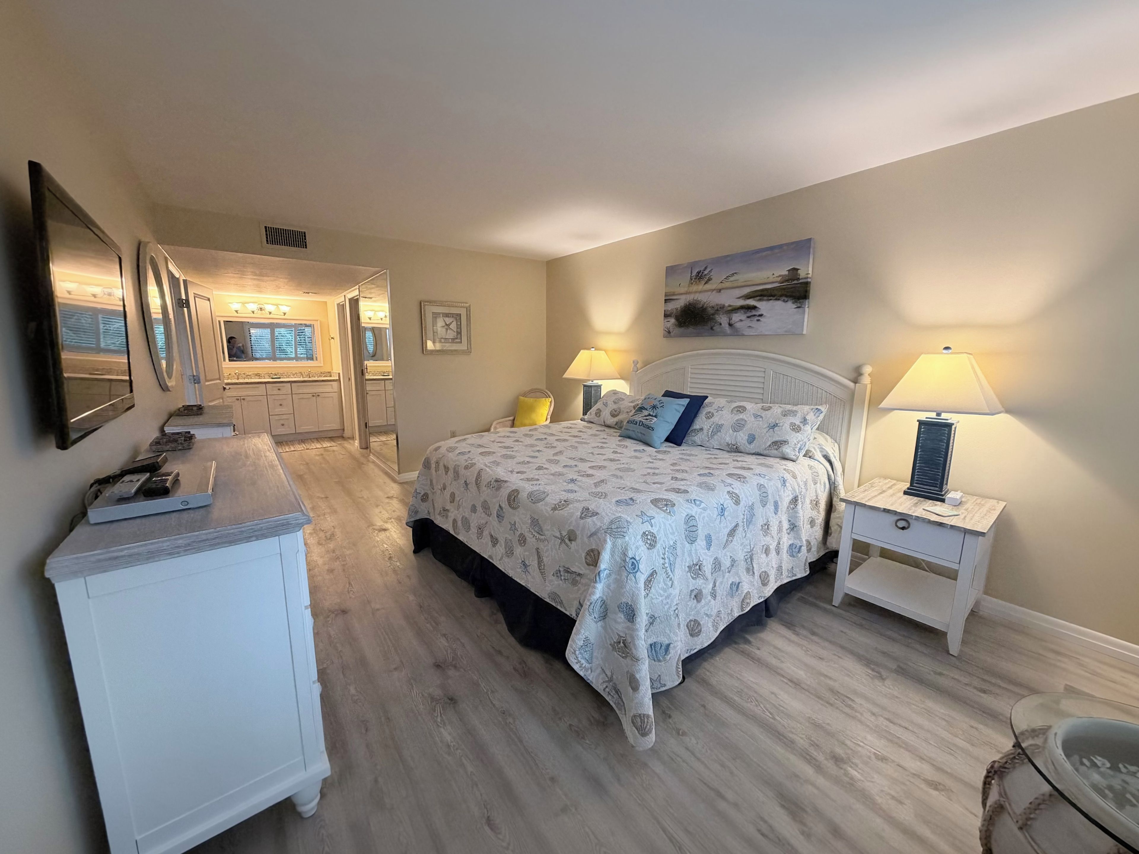 Un dormitorio con cama, mesitas de noche, cómoda, espejo y un cuadro en la pared.
