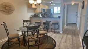 A dining area with a glass table and metal chairs, a kitchen in the background, and a hallway leading to another room. - Siesta Dunes 5-303, Come enjoy our little piece of Paradise! (Siesta Key)