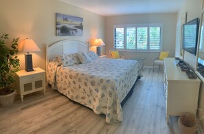 A bedroom with a bed, bedside lamps, a chair, a desk, a window with blinds, and a potted plant.