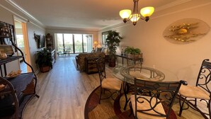 A spacious living area with a glass dining table surrounded by chairs, a sofa, and a coffee table. There is a chandelier above the table, and a large window provides natural light. - Siesta Dunes 5-303, Come enjoy our little piece of Paradise! (Siesta Key)