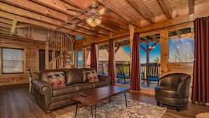 A cozy cabin interior with a wooden ceiling, a leather sofa with patterned cushions, a coffee table, and a view of the outside through large windows. - 🌙 Midnight Wonder 🏔️ Twinkling Valley Lights, Hot Tub & Resort Pool (Sevierville)