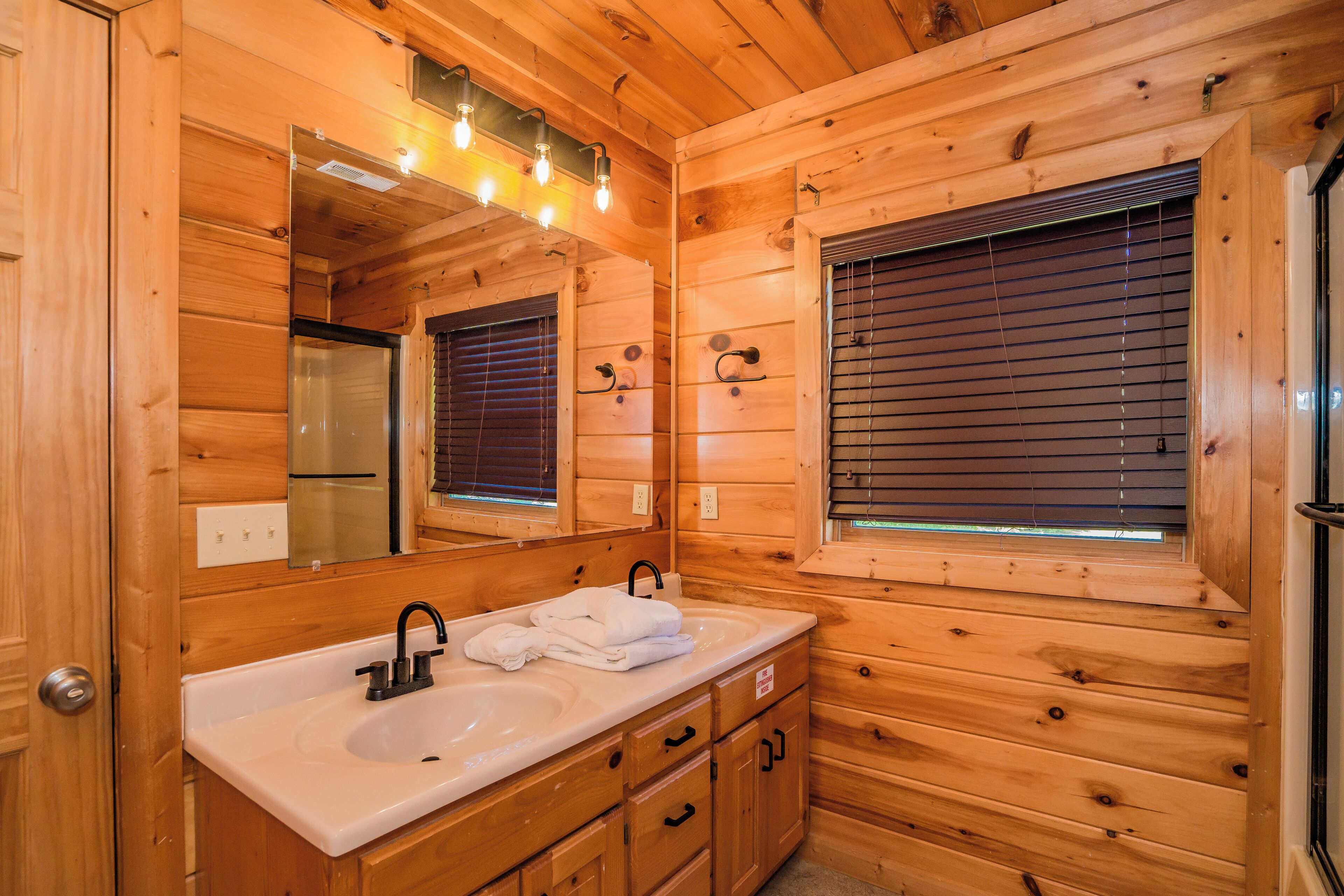 A wooden cabin bathroom with a double sink, a mirror, and a window with blinds.
