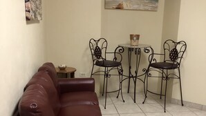 Un espace de vie comprenant un canapé en cuir marron, une petite table ronde avec un panier, et deux chaises en métal dotées de dossiers décoratifs. Un tableau représentant une scène de plage est accroché au mur, au-dessus de la table.