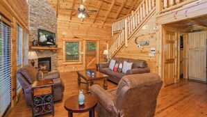 A log cabin interior with a stone fireplace, leather sofas, a wooden staircase, and a television mounted on the wall. - Bearly Wet: Indoor Pool, Mtn Views! Hot tub, Wifi, Game Room, near attractions (Sevierville)