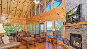A spacious living room with wooden floors, leather furniture, a stone fireplace, and large windows. - Bearly Wet: Indoor Pool, Mtn Views! Hot tub, Wifi, Game Room, near attractions (Sevierville)