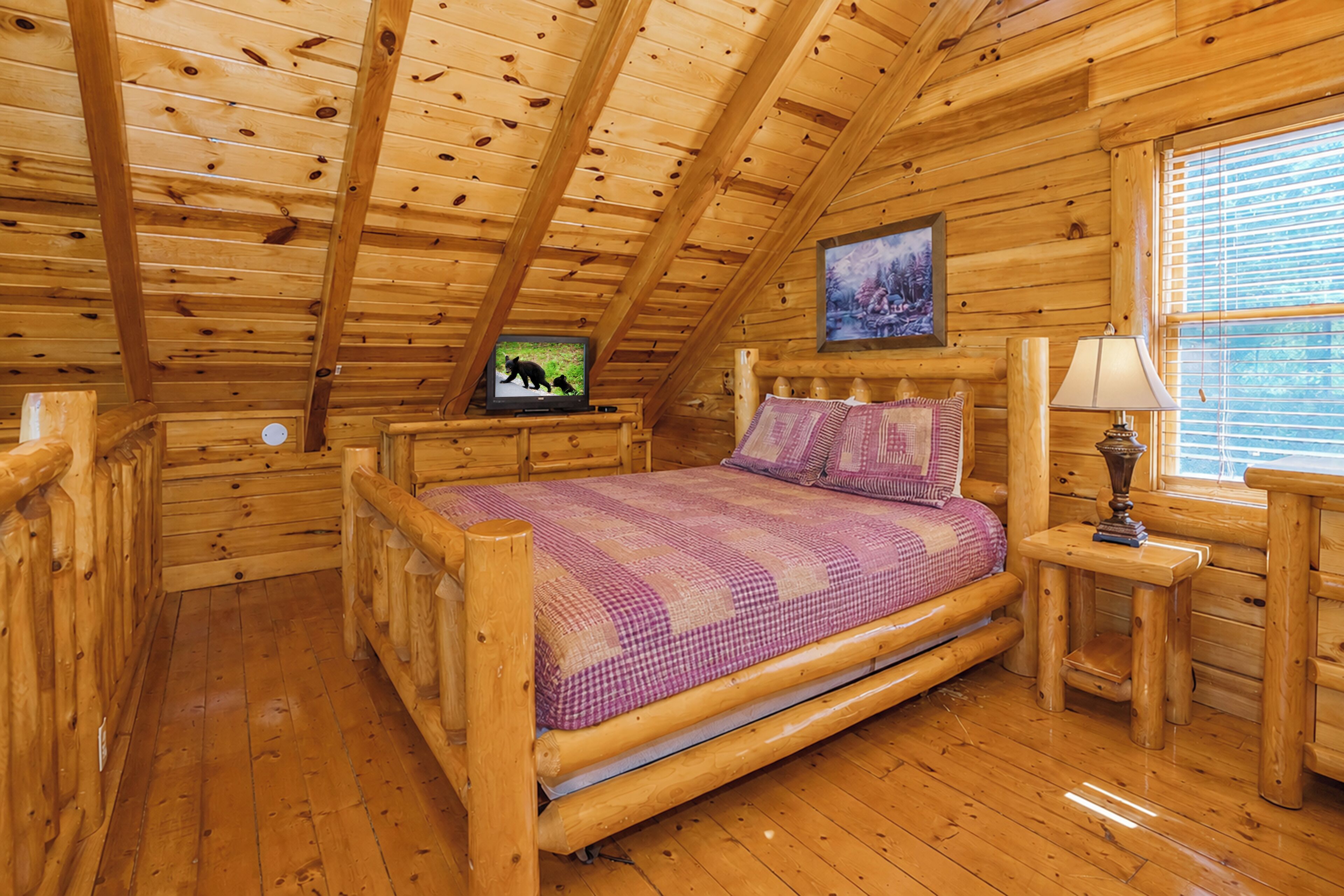 Une chambre située dans un chalet en rondins, avec un lit en bois, un téléviseur posé sur un meuble, une fenêtre avec des stores et une lampe sur une table de chevet.