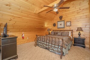 A bedroom with a wooden ceiling, a bed with patterned bedding, a nightstand with a lamp, and a dresser.