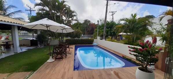 Ein Garten mit Swimmingpool, Holzterrasse, Gartenmöbeln und einem großen Sonnenschirm.