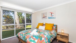 Ein Schlafzimmer mit großem Fenster, einem Bett mit farbenfroher Bettwäsche, zwei Kissen, einem Nachttisch mit Lampe und zwei Wandbildern.