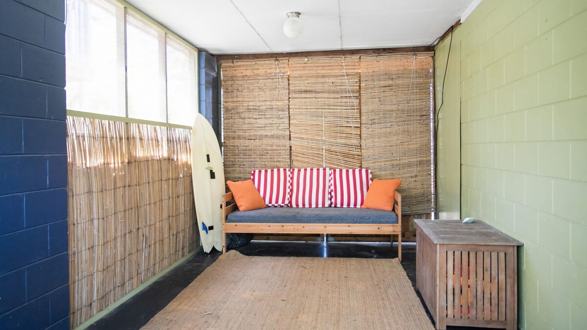 A small room with a bamboo wall, a bed with striped pillows, and a surfboard leaning against the wall.