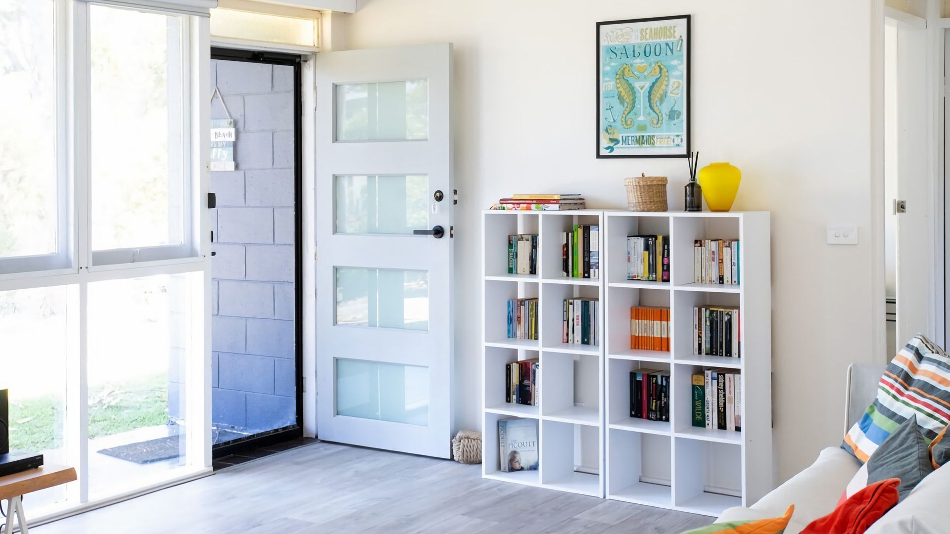 Ein heller Raum mit einer weißen Tür, einem Bücherregal voller Bücher und einem gerahmten Poster an der Wand.