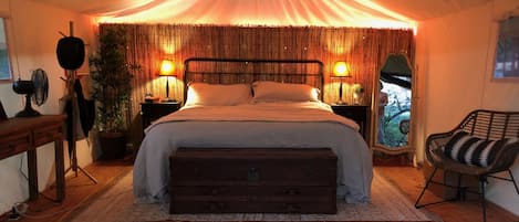 A tented room with a bed, bedside lamps, a wooden bench, a chair, and a view of the outdoors.