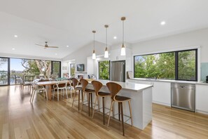A modern kitchen with a central island, bar stools, and a dining area with a view. - Alto Vista (Lorne)