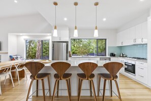 Une cuisine moderne avec des meubles blancs, un réfrigérateur en acier inoxydable et une cuisinière à gaz. On y trouve trois tabourets de bar en bois et une table à manger avec des chaises.