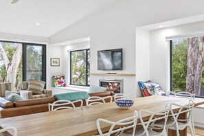 A modern living room with a wooden dining table, white chairs, a sofa with patterned cushions, a fireplace, and a television mounted on the wall. - Alto Vista (Lorne)