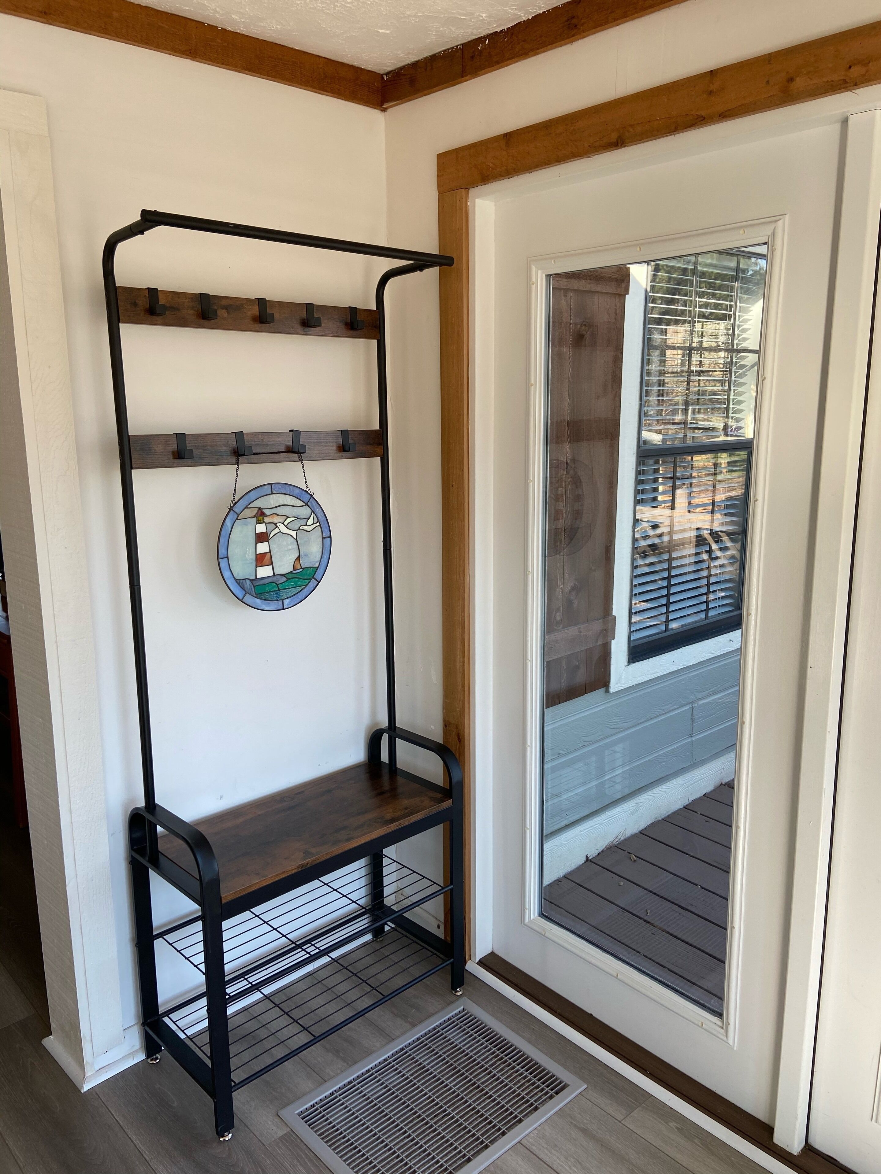 Un coin avec une étagère en métal noir adossée à un mur blanc, un petit banc en bois et une porte avec une fenêtre.