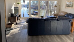 Een woonkamer met een grote, donkere bank, een televisie op een houten dressoir en uitzicht op een steiger met boten door grote ramen.