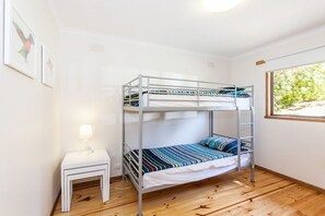 Ein Schlafraum mit Etagenbetten, einem kleinen weißen Tisch und einer Lampe, ein Fenster mit Blick auf Grünflächen und eine Wand mit gerahmten Kunstwerken.