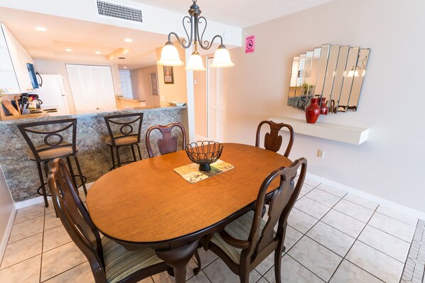 A dining area with a wooden table and chairs, a chandelier, a kitchen in the background, and a mirror on the wall. - Oceanfront 2BR Condo 6th Floor Balcony with Stunning Ocean Views, 603 (Indian Harbour Beach)