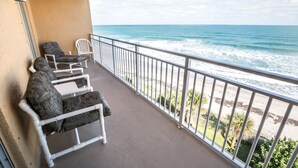 Un balcone con due sedie bianche e un cuscino decorato, con vista su una spiaggia, l'oceano e un cielo sereno.