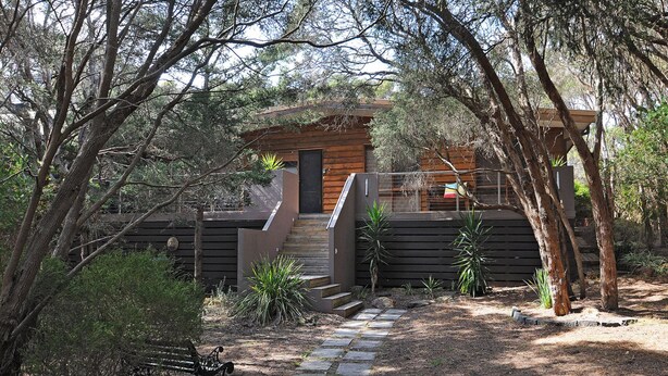 Une cabane moderne avec une façade en bois, un chemin de pierre menant à l’entrée et un banc à proximité.