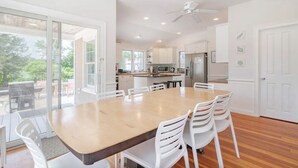 A modern kitchen with white cabinets, a stainless steel refrigerator, and a dining area with a wooden table and white chairs. - Walk to beaches & ferries ★ Snow's Creek Waterview (Barnstable)